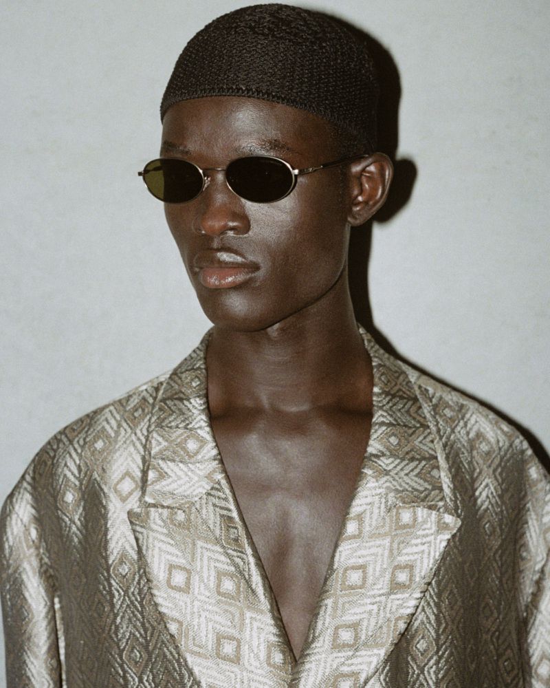 Close-up studio shot of a dark-skinned adult male in a patterned jacket and dark beanie, wearing oval sunglasses against a light gray backdrop.