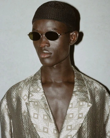 Close-up studio shot of a dark-skinned adult male in a patterned jacket and dark beanie, wearing oval sunglasses against a light gray backdrop.