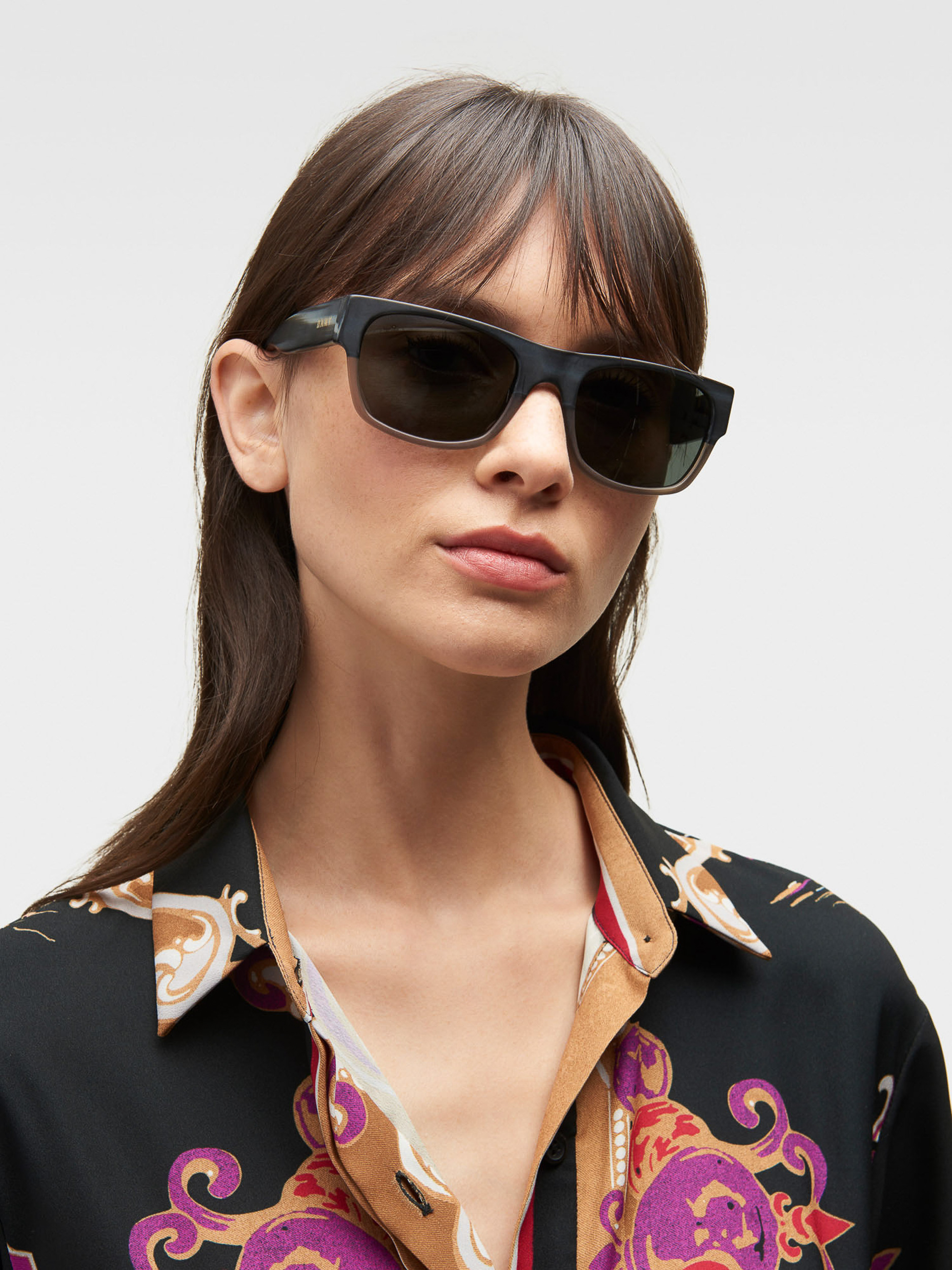 Woman wearing sunglasses and a patterned black, beige, and red collared shirt. Dark hair, studio lighting, and white background.