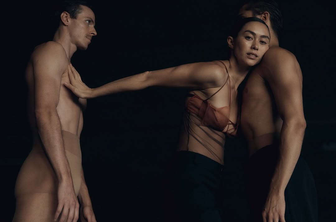 Three figures, two adult males and one adult female, are in a close pose in a dimly lit indoor studio environment.