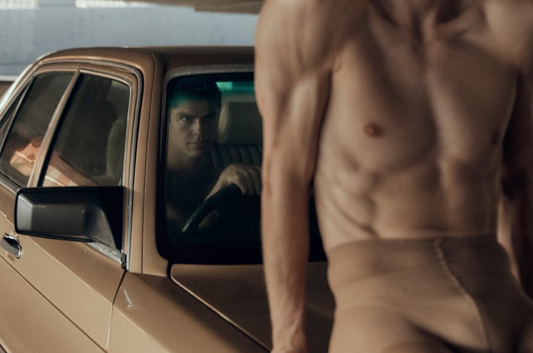 Tan car interior features a man driving while another shirtless man in tights stands outside, showcasing athletic physique and neutral tones.