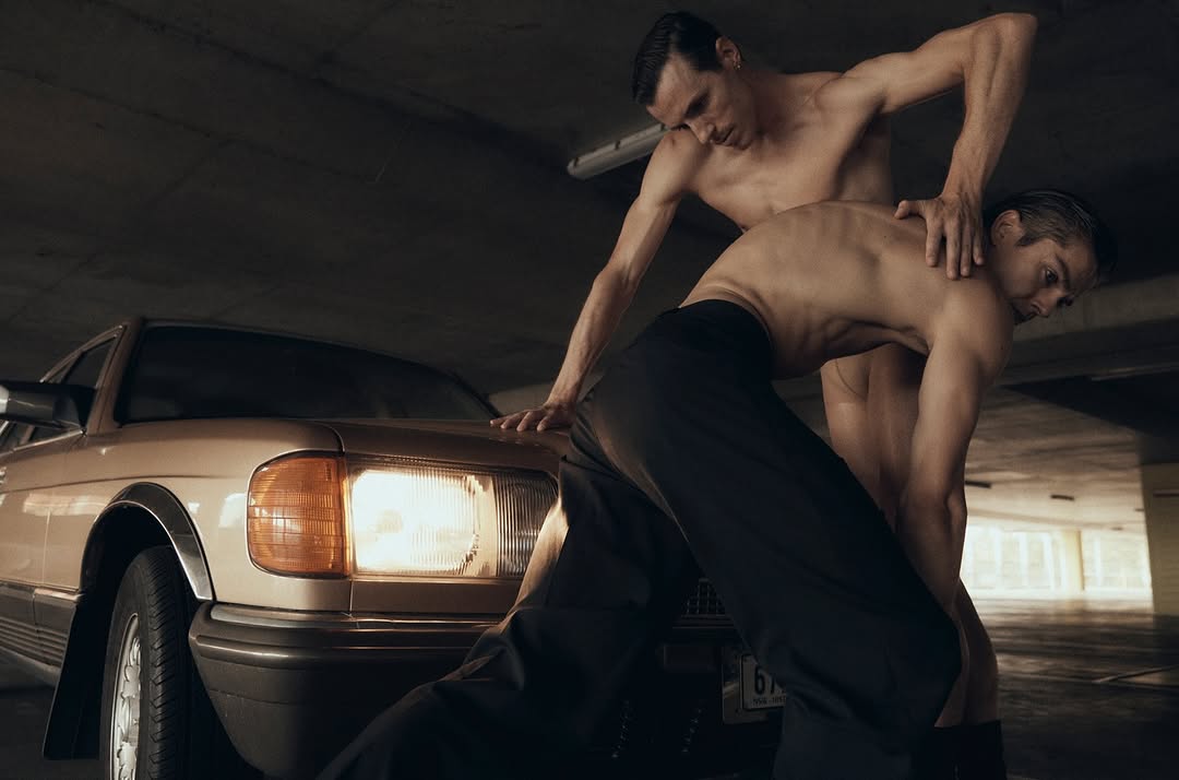 Two shirtless adult males pose with a tan classic car inside an indoor parking garage with overhead fluorescent lighting. Moody automotive style.