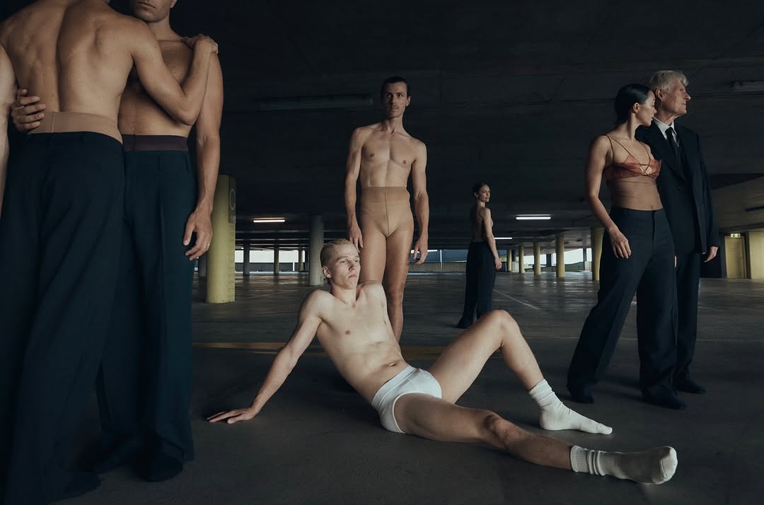 Artistic shot of diverse group of people in an underground parking lot; some shirtless, some in business suits, and some in dancewear.