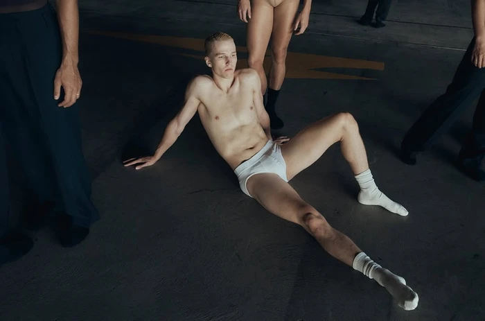 Blonde man in white briefs seated on a concrete floor surrounded by standing people in an industrial space