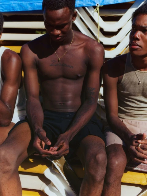 Three young Black men in tank tops and shorts are sitting outside in sunlight, with folded white deck chairs in the background.