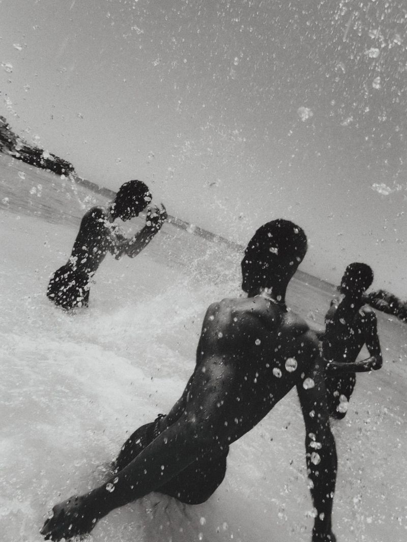 Monochrome, low-angle, medium shot of three dark-skinned people in a body of water with splattering droplets in foreground, and a landmass in background.