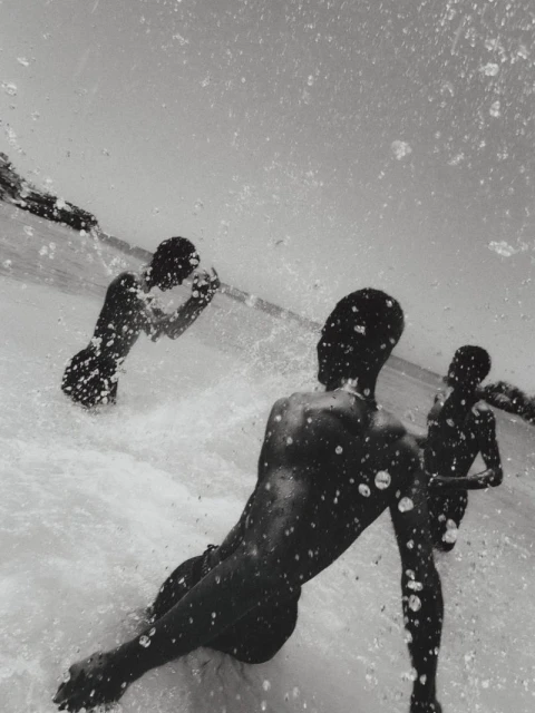 Monochrome, low-angle, medium shot of three dark-skinned people in a body of water with splattering droplets in foreground, and a landmass in background.