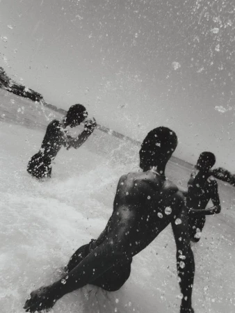 Monochrome, low-angle, medium shot of three dark-skinned people in a body of water with splattering droplets in foreground, and a landmass in background.