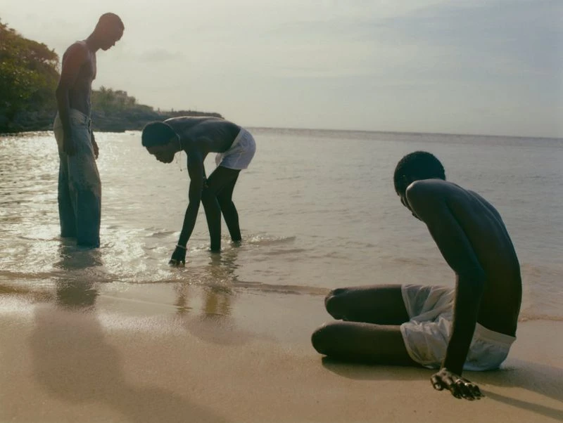 Three Black men on a beach: one standing in the water, one bending over, and one sitting on the sand, at ocean's edge.