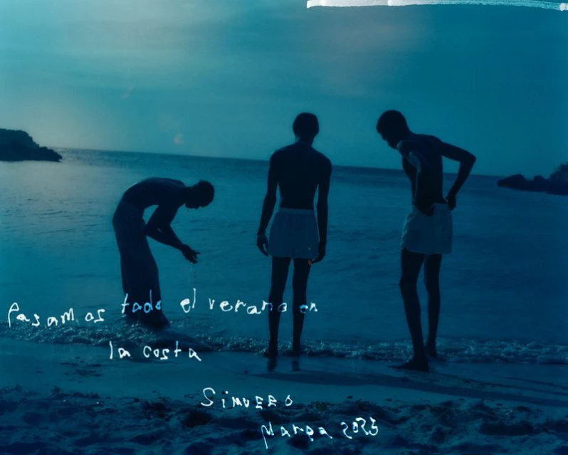 Three young adult males gather at dusk on a beach with gentle waves; one male in swimwear bends over, and two stand looking at the water.