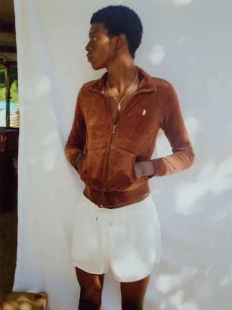 An African American adult male poses in a brown jacket and white shorts against a neutral background, shot in natural lighting.