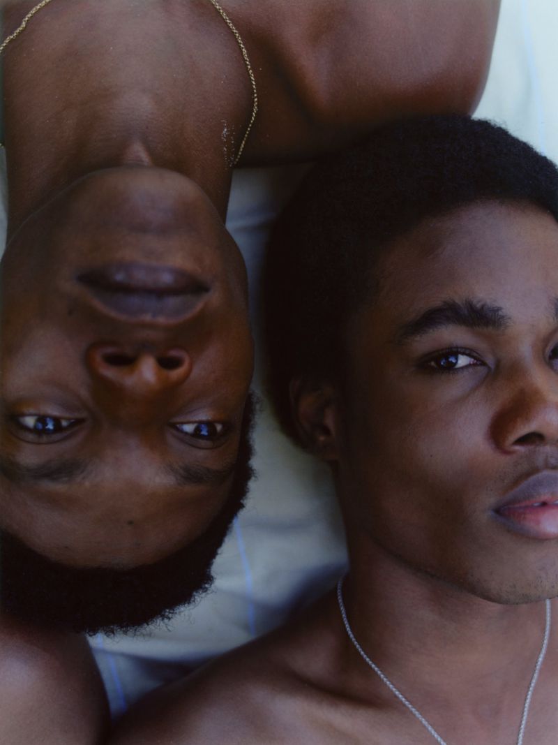 Portrait of two young adult African American men lying side by side; they both wear silver chain necklaces indoors with natural lighting.