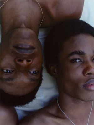 Two young Black men lying head-to-shoulder wearing delicate chain necklaces against pale fabric in a close-up portrait.