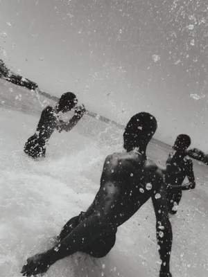 Black and white photograph of three figures swimming and moving through ocean surf with water droplets splashing across the frame.