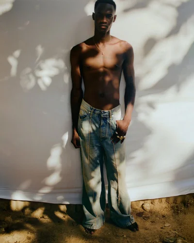 Shirtless black man wearing light denim jeans standing against a plain white backdrop with dappled shadows