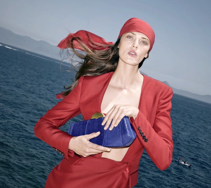 Fashion shot of a woman in a red suit holding a blue purse with a blue ocean and mountains in the background on a sunny day.