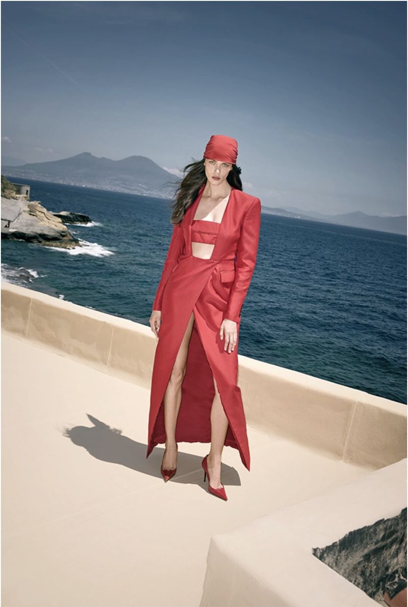 A woman in a red outfit stands on a rooftop with the ocean and a mountain in the background, creating a fashionable and scenic portrait.