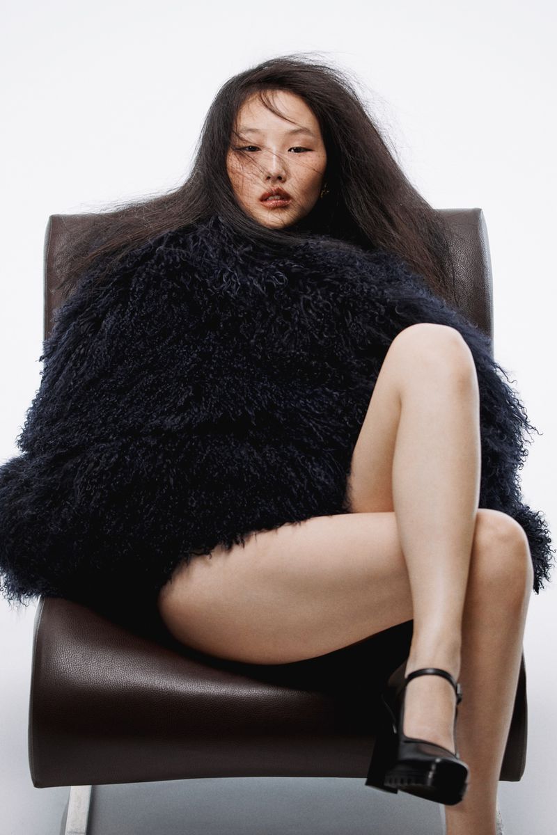 Attractive Asian woman with long black hair sits on a leather chair in a photo studio wearing a large black fur jacket and black high heels.