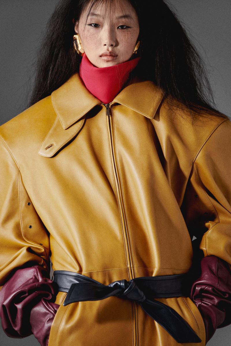 Eye-level shot of Asian woman in a yellow leather coat, red turtleneck, and large gold earrings posing in a studio with a grey background.