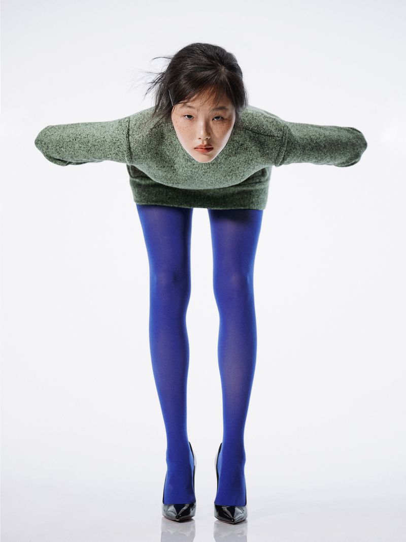 An adult woman in a bent-over pose, wearing a green sweater, blue tights, and black high-heeled shoes against a white backdrop.