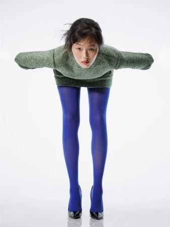 An adult woman in a bent-over pose, wearing a green sweater, blue tights, and black high-heeled shoes against a white backdrop.