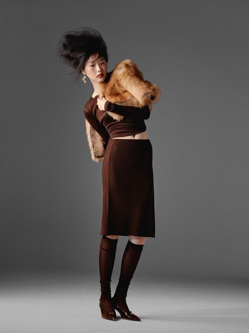 Elegant Asian woman in studio wearing brown dress, knee-high socks, and fur stole. Dark hair blown back, neutral background, full shot.