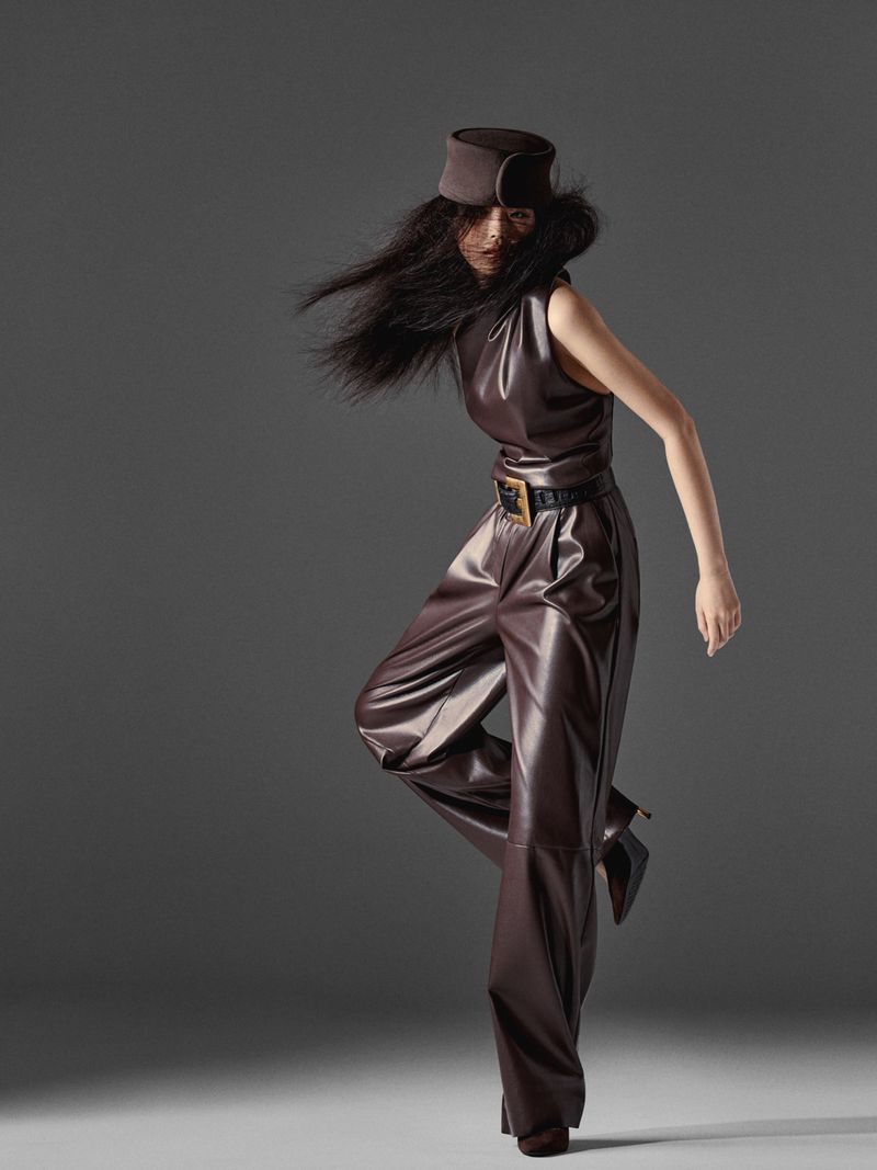 Model Peng Chang wearing a dark brown leather jumpsuit and matching sculpted hat in a dynamic pose against a grey studio backdrop.