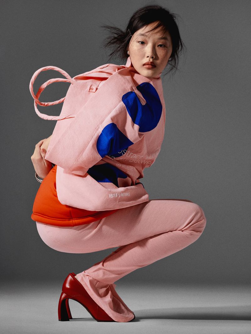 Fashion model poses in a studio wearing a pink hoodie and leggings, paired with red high-heeled shoes against a grey background.