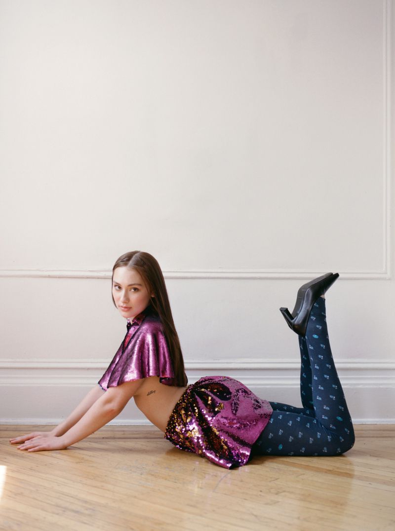Woman in shimmering purple cape and skirt, dark patterned tights and heels, lying on stomach on a wood floor against a white wall.