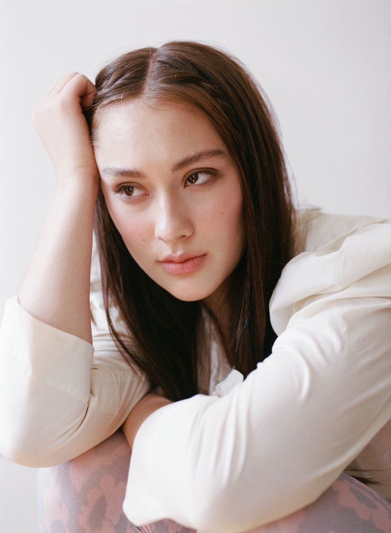 Portrait of a young woman with dark hair and light skin, posed indoors against a white background. She wears a white blouse.