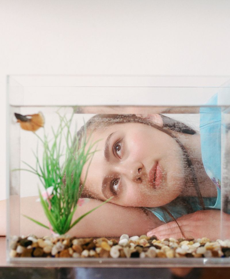 Teen girl rests near a fishbowl with a betta fish and artificial plant. Indoor scene with white wall backdrop and subdued lighting.