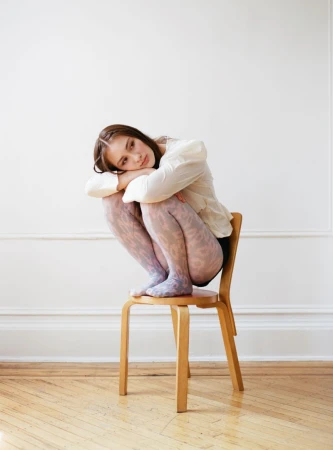Lola Tung crouching on a wooden chair wearing a white blouse and patterned tights against a white wall with wood flooring.