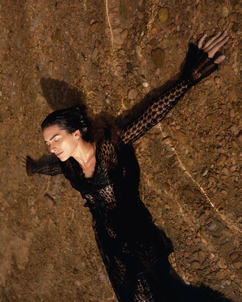 Woman in a black lace dress lying in shallow water with rocks, creating a serene and ethereal scene with soft, diffused lighting.