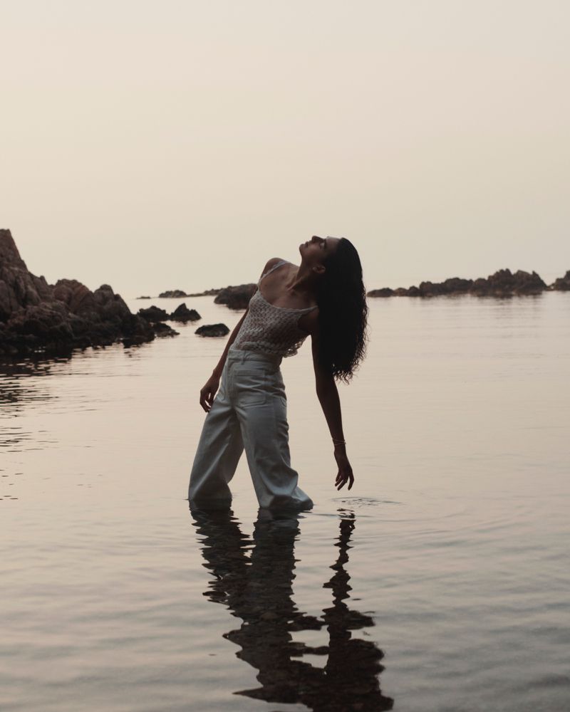 A woman in a tank top and pants stands in shallow water near rock formations at dusk, creating a serene and reflective scene.