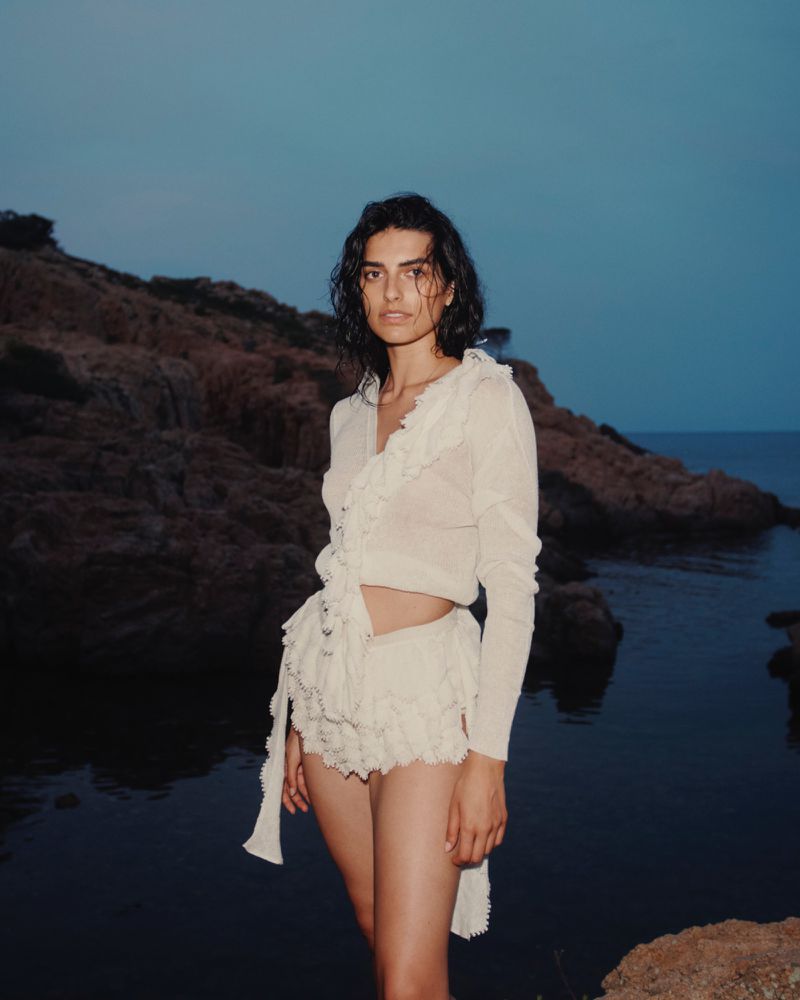 Portrait of a woman in ruffled white outfit standing on the coast with a dark ocean and rock formations under a dim blue sky at dusk.