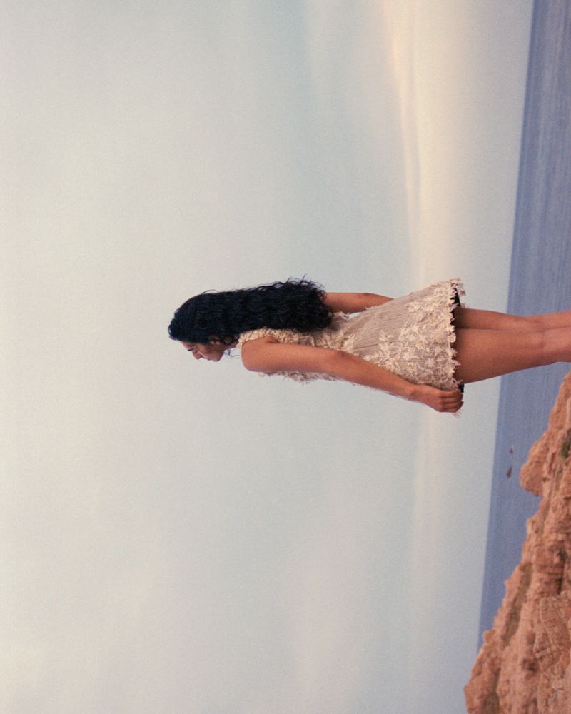 Woman in a white dress stands on a rocky cliff overlooking the ocean, sky is visible, creating a scenic and elegant portrait.