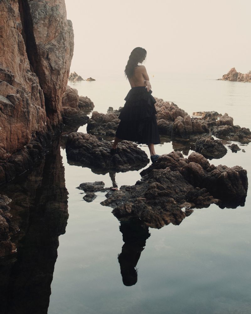 Woman in black dress standing on rocks by the water's edge, with her reflection visible in the water, and coastal rock formations.