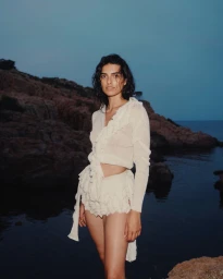 Model wearing a white ruffled top and shorts standing on a rocky coast near the water at dusk.