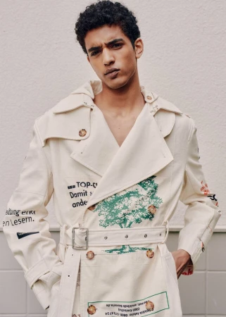 Portrait of a young adult male model with dark hair wearing a printed white trench coat, posing against a neutral background.