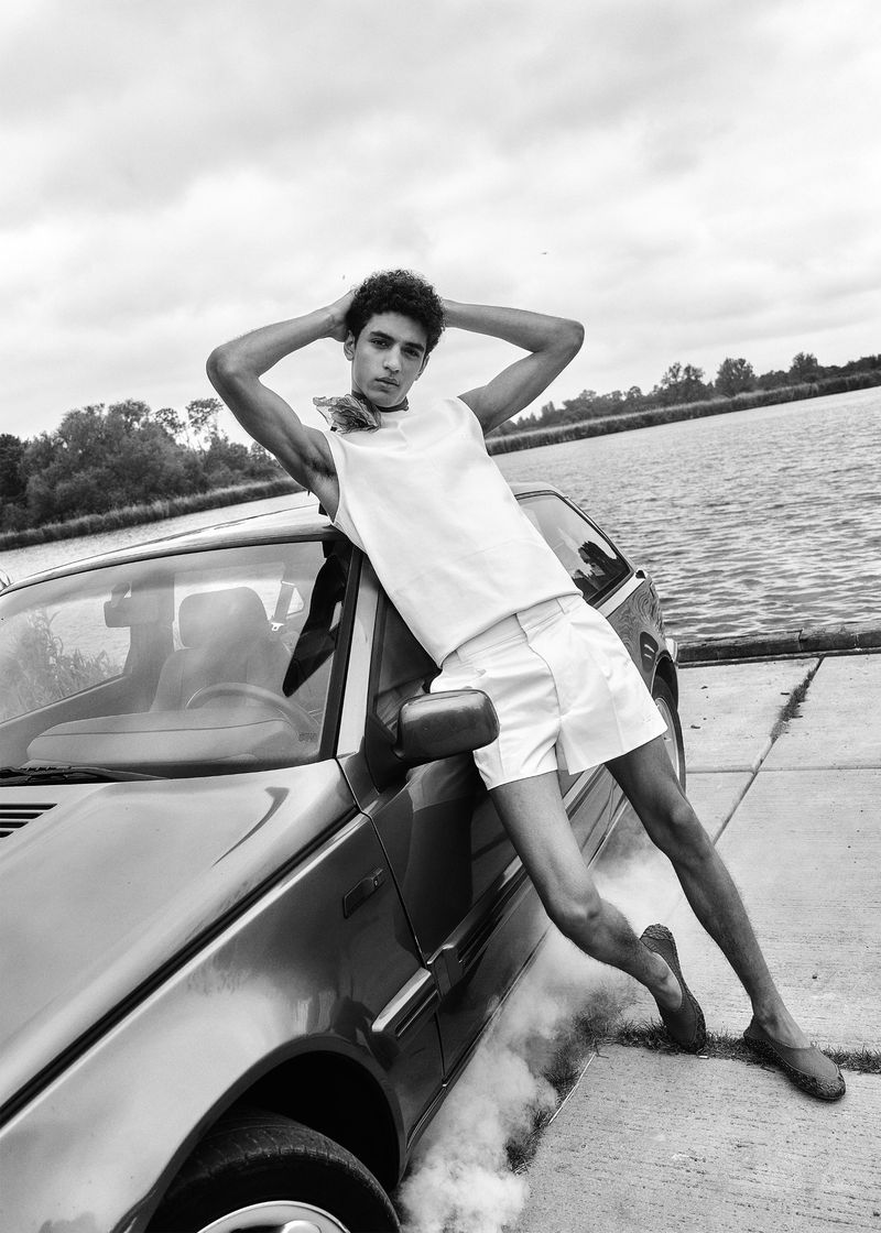 Black and white portrait of a young man in a white top and shorts leaning against a car, with a lake and trees in the background.