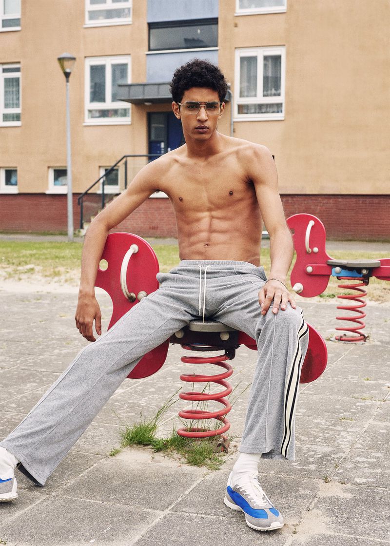 Athletic man with six-pack abs sits topless on a red playground spring rider with gray sweatpants, wearing sneakers, eyeglasses, outdoors.