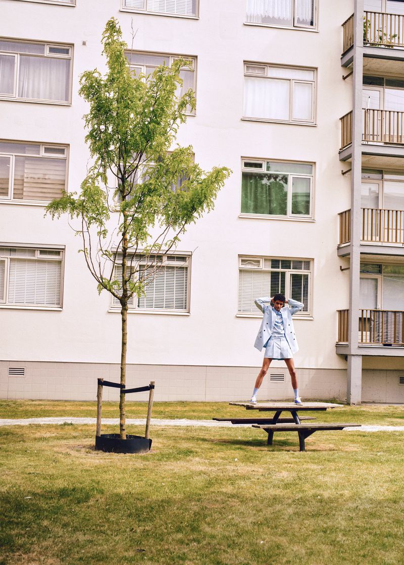 A woman in a light blue suit is standing on a picnic table next to a small tree, in front of a white apartment building on a lawn.