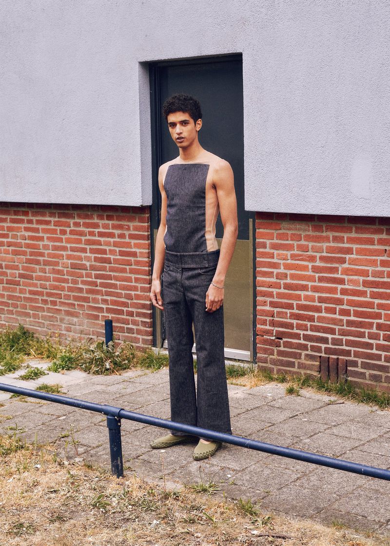 Man poses in open-backed denim jumpsuit and sage flats against a brick and concrete wall outdoors on a sunny day.