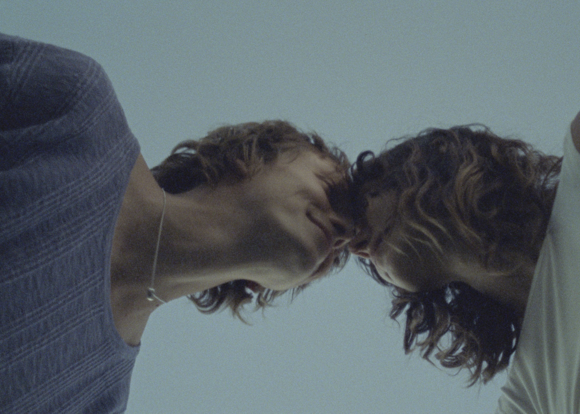 Intimate low-angle shot of a young adult couple with curly hair, facing each other nose-to-nose. A simple pendant necklace is visible on one person.