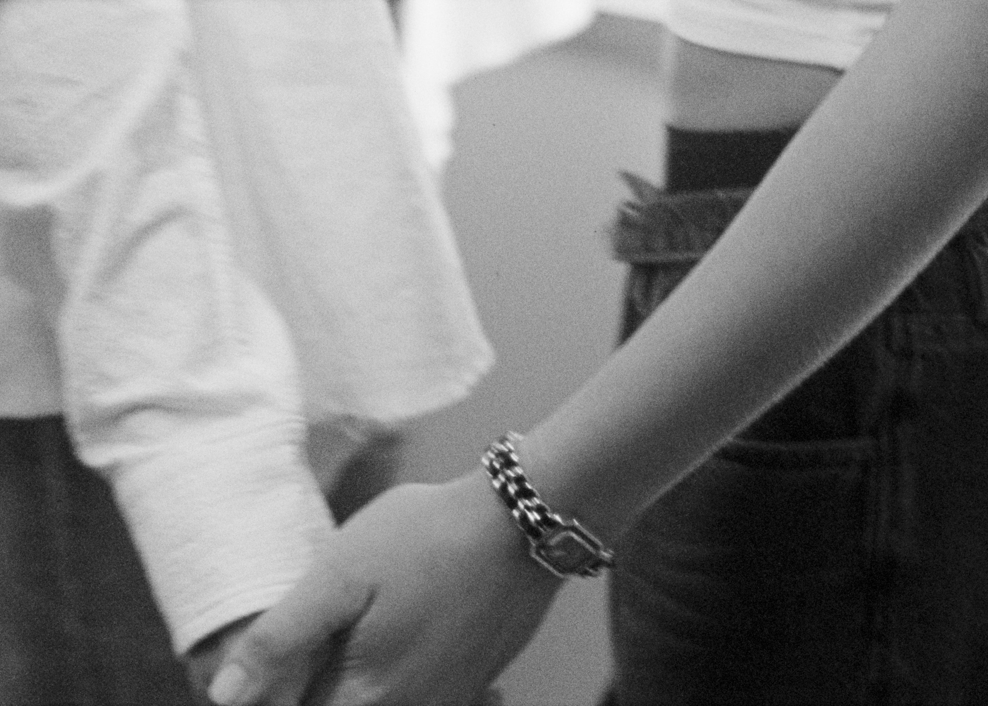 Close-up black and white shot of two people holding hands. One person wears a bracelet and jeans, the other wears a white long-sleeve shirt.