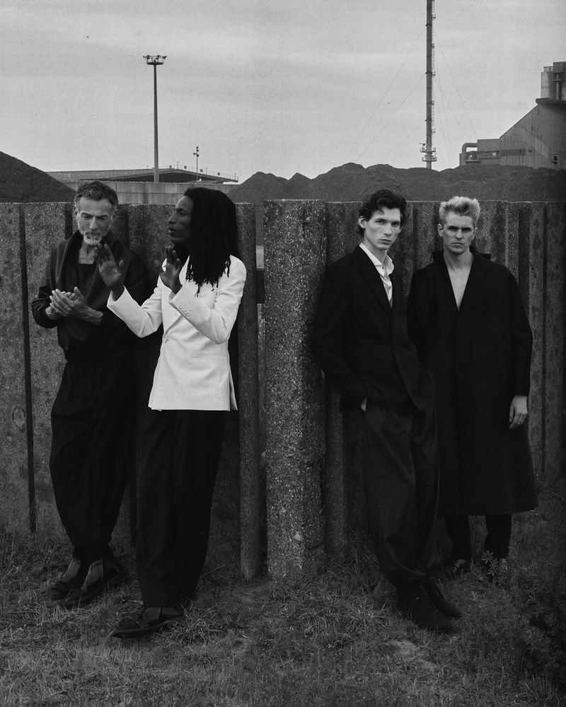 Four people posing in a black-and-white fashion editorial shot against a concrete wall with industrial background elements.