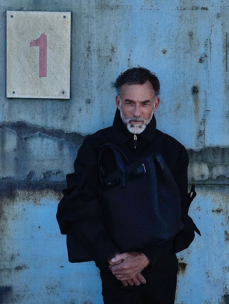Mature man in black jacket and slacks poses with a tote bag against a distressed wall next to a numbered sign, capturing a street-style vibe.
