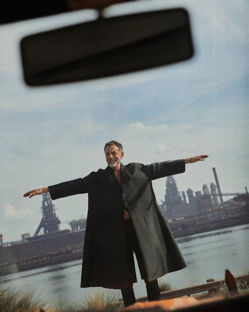 Mature man in black coat stands with arms outstretched near water, old industrial buildings, and cloudy sky, creating dramatic image.