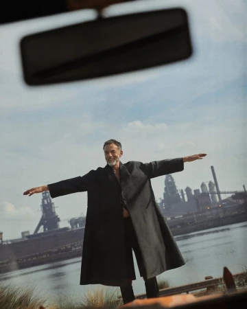 Mature man in black coat stands with arms outstretched near water, old industrial buildings, and cloudy sky, creating dramatic image.