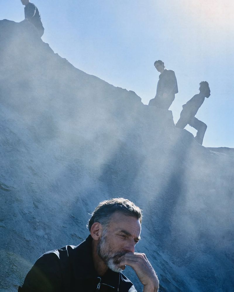 Pensive man with a beard rests his chin on his hand while seated near a misty landscape with silhouetted figures climbing the slope behind.